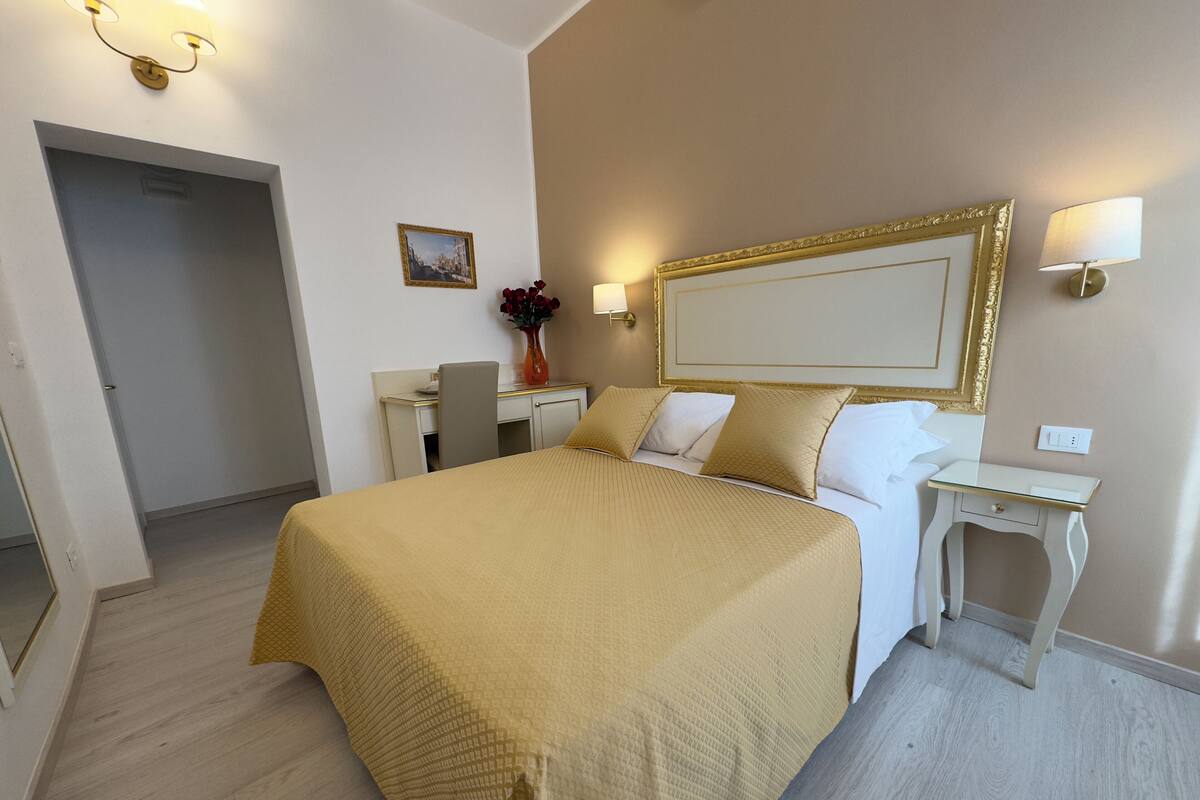 A hotel room with a large bed, a desk, a chair, a small table, and a wall-mounted lamp.