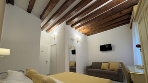 A bedroom with a bed, a sofa, a desk, a TV, and wooden beams on the ceiling. - Residenza Briati (Venice)