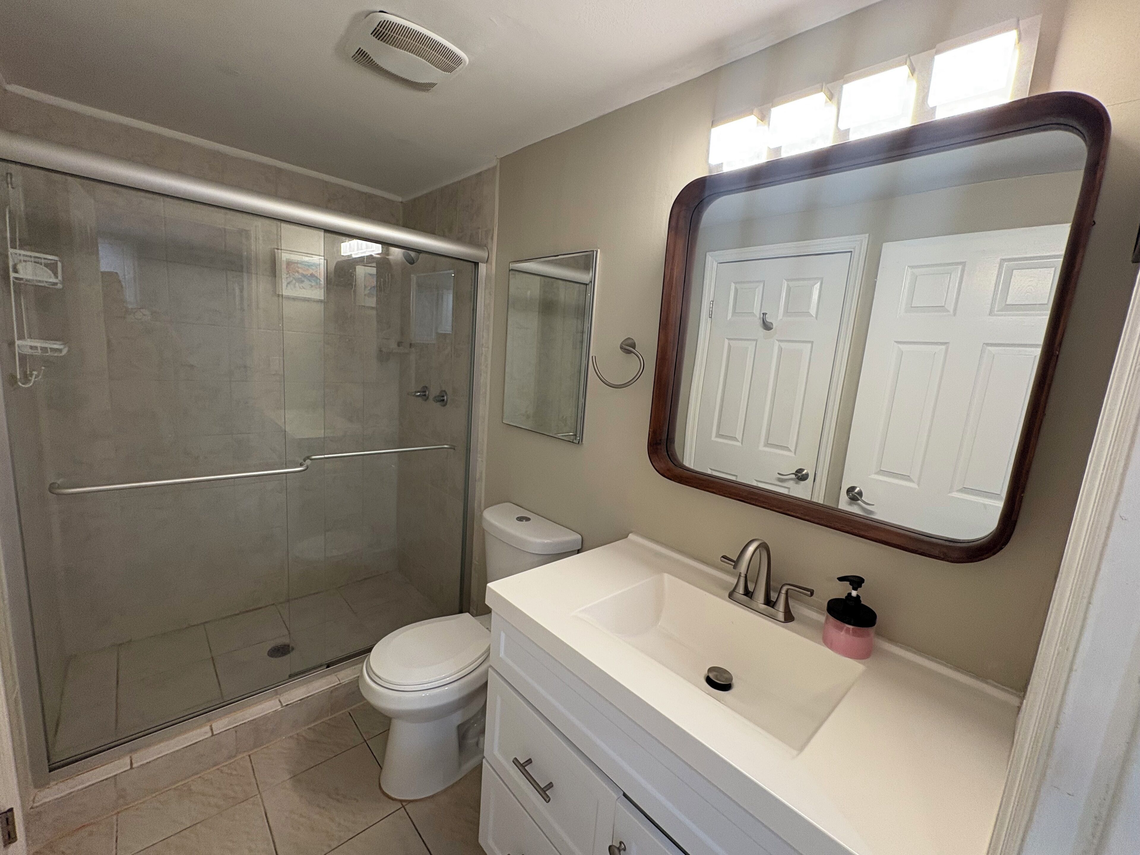 A bathroom with a glass-enclosed shower, a toilet, a sink with a mirror above it, and a vanity with drawers.
