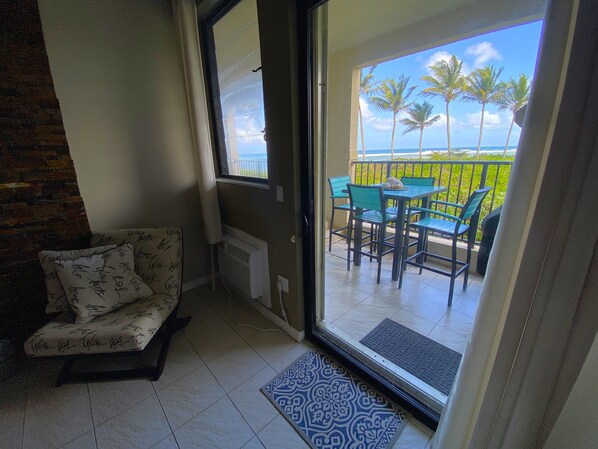 En balkon med bord og stole, en sofa og havudsigt med palmer.