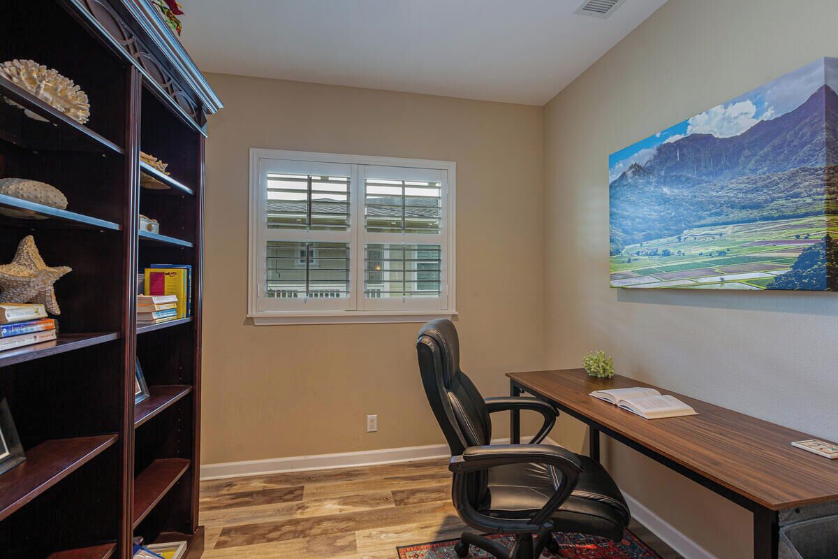 A room with a wooden desk, a black office chair, a bookshelf with various items, a window with blinds, and a landscape painting on the wall.
