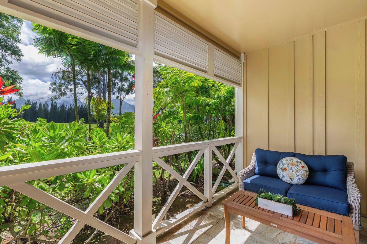 A covered outdoor seating area with a blue sofa and wooden table, surrounded by lush greenery and a view of mountains.
