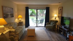Sala de estar con sofá, sillones, televisión y un área de comedor visible a través de un vidrio.