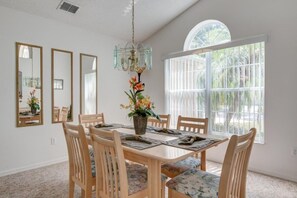A dining area with a wooden table, six chairs, a chandelier, and a large window with a view of greenery. - Perfect Family Home Pool & Spa! 4 Bedroom Home by RedAwning (Haines City)