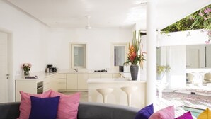 A modern living room with a grey sofa, white kitchen, and a pool area visible through an open space. - Villa La Sirena 2 Minutes From Beach 3 Bedroom Villa by RedAwning (Seminyak)