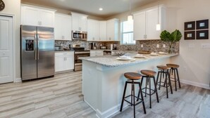 A modern kitchen with white cabinets, a large island with stools, stainless steel appliances, and a stone backsplash. - Champions Gate 6bed 5bath Vacation Home 6 Bedroom Villa by RedAwning (Davenport)