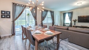 A dining area with a wooden table set for four, a sofa, and a TV mounted on the wall. - Champions Gate 6bed 5bath Vacation Home 6 Bedroom Villa by RedAwning (Davenport)