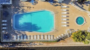 Eine Vogelperspektive zeigt einen Poolbereich mit einem großen rechteckigen Pool, einem kleineren kreisförmigen Pool und mehreren Liegestühlen, die um die Pools herum platziert sind.