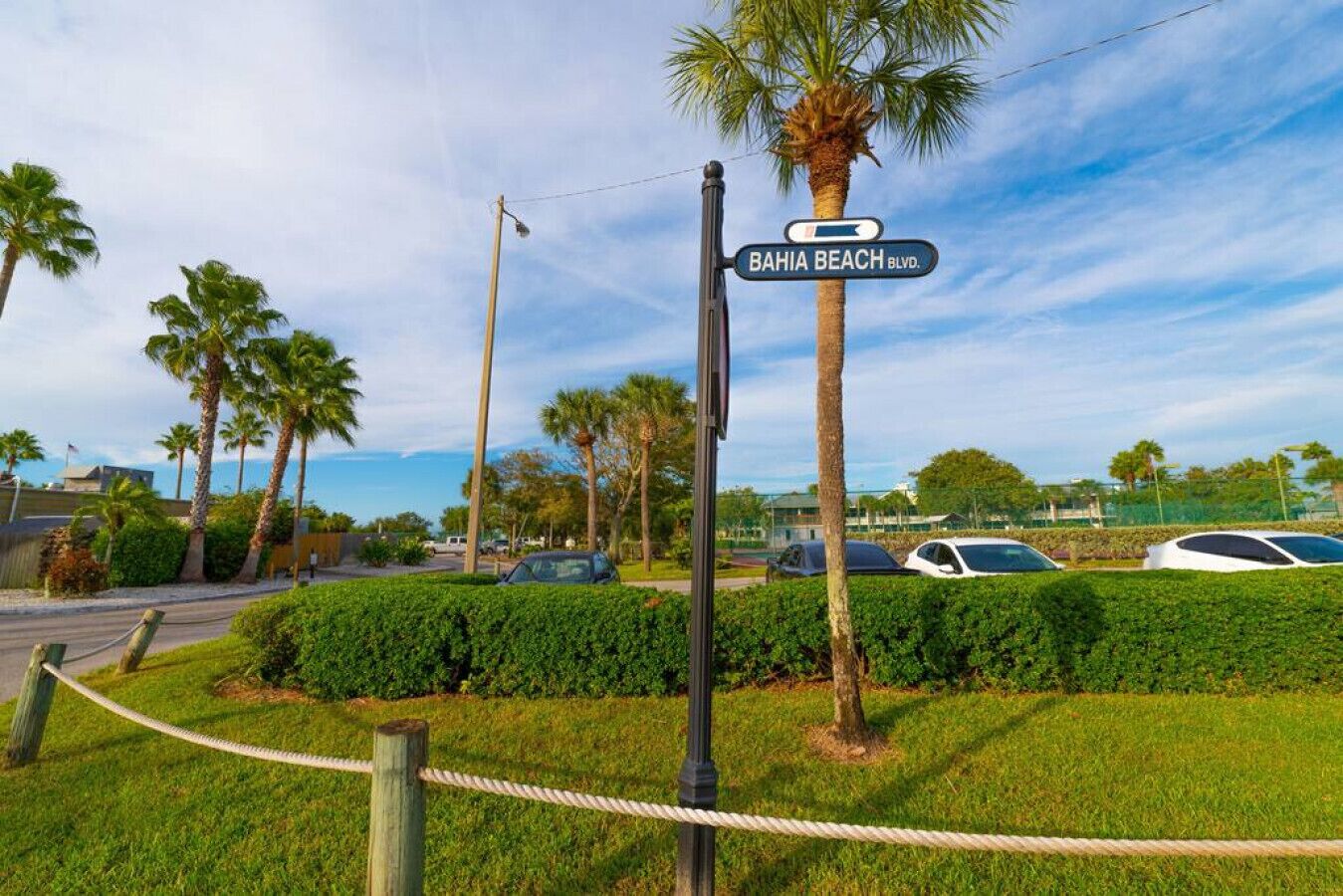 Ein Straßenschild für die 'Bahia Beach Blvd.' mit blauem Himmel, Palmen und geparkten Autos.