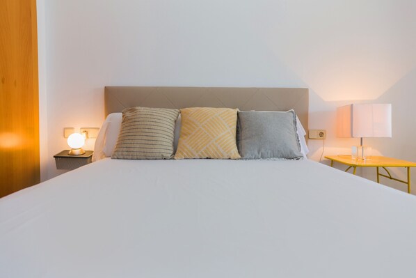 A neatly made bed with three pillows, a bedside table with a lamp, and a small table with a lamp. - Golden Gardens Saudade Apartment Javea Arenal (Javea / Xàbia)