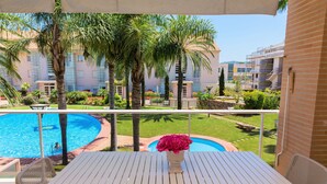 Un balcon avec une table et des chaises blanches, donnant sur une piscine et un jardin aménagé avec des palmiers.