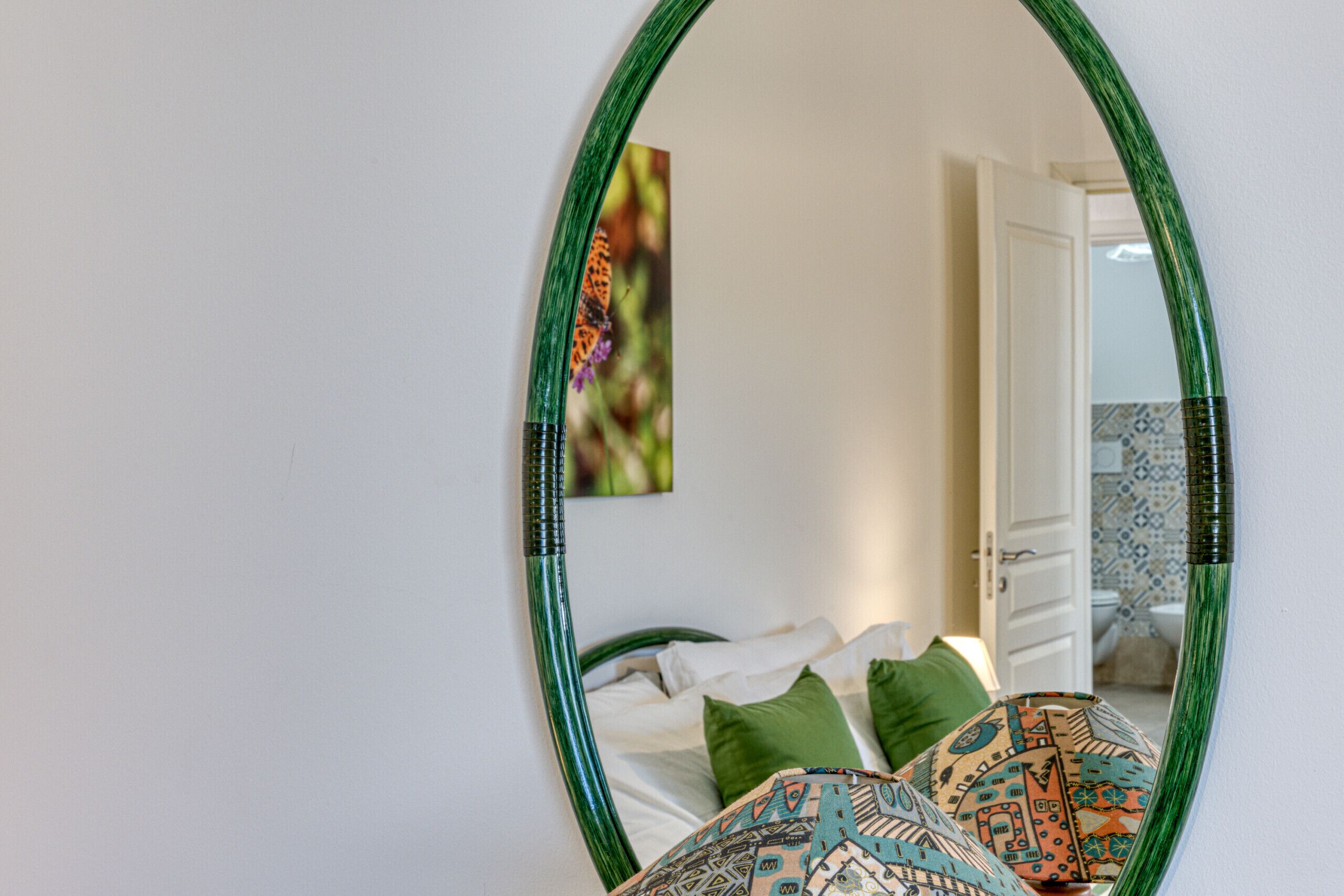 Ein Schlafzimmer mit Bett, Kissen und einem spiegelnden, grün gerahmten Spiegel, der eine Tür und ein Badezimmer zeigt.