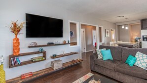 Un soggiorno moderno con una TV a schermo piatto, mensole in legno con oggetti decorativi, un divano grigio con cuscini fantasia e un'area cucina sullo sfondo.