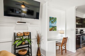 Una sala de estar moderna con un televisor de pantalla plana, un cuadro de una escena tropical, una silla de madera, una mesa de vidrio y una zona de cocina al fondo.