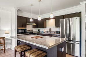 A modern kitchen with a central island, stainless steel appliances, dark cabinetry, and pendant lighting. - Koa Resort 1B: Garden View Condo w/ AC, Shared Hot Tub & Pool (Kihei)