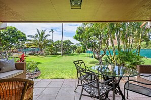 A covered patio area with a glass table and chairs, a wicker sofa, and a view of a well-maintained lawn and trees. - Koa Resort 1B: Garden View Condo w/ AC, Shared Hot Tub & Pool (Kihei)