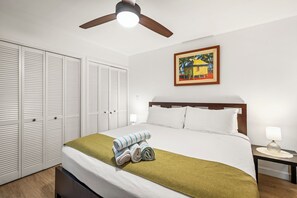 Habitación de hotel con cama, mesita de noche, lámpara, ventilador de techo y un cuadro en la pared.