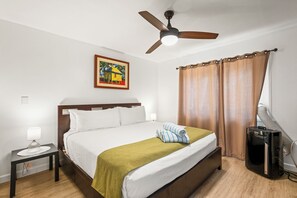 Habitación de hotel con cama, ventilador de techo, un cuadro, ventana con cortinas, mesita de noche y cafetera.