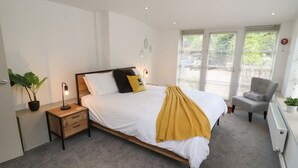 A modern bedroom with a bed, bedside table, lamp, chair, and a window with blinds. - The Coach House (Ross-on-Wye)