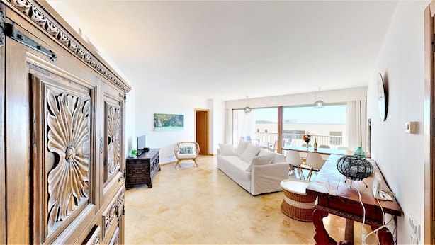 A spacious living room with a white sofa, a wooden cabinet with intricate carvings, a wicker chair, and a dining area with a table and chairs. - Lomas de los Monteros - Apartment with seaviews (Marbella)