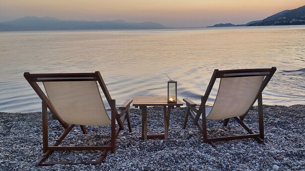 Two wooden chairs and a table on a pebbled beach with a view of calm water and distant mountains.