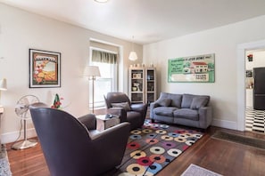Een woonkamer met een bank, fauteuil en een ventilator. Er is een raam met uitzicht, een lamp en een poster aan de muur.