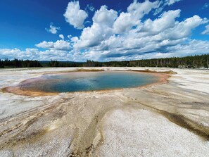 A large, circular geothermal pool surrounded by a barren landscape with sparse vegetation and a forest in the background. - Moose Willow at Teton Valley Jackson Hole (Victor)