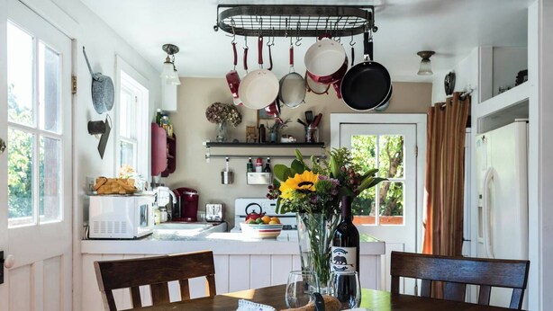 Une cuisine avec des casseroles suspendues, une table à manger avec du pain et du fromage, et un vase rempli de fleurs.