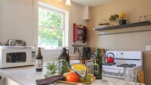 Une cuisine équipée d’un four à micro-ondes, d’une cuisinière, et sur le plan de travail, on peut voir diverses bouteilles et des légumes.