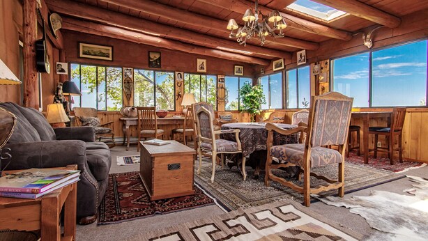 A rustic cabin interior with wooden walls, a chandelier, a dining table, and a large window offering a view of trees. - Rustic Ranch Retreat (Lamy)