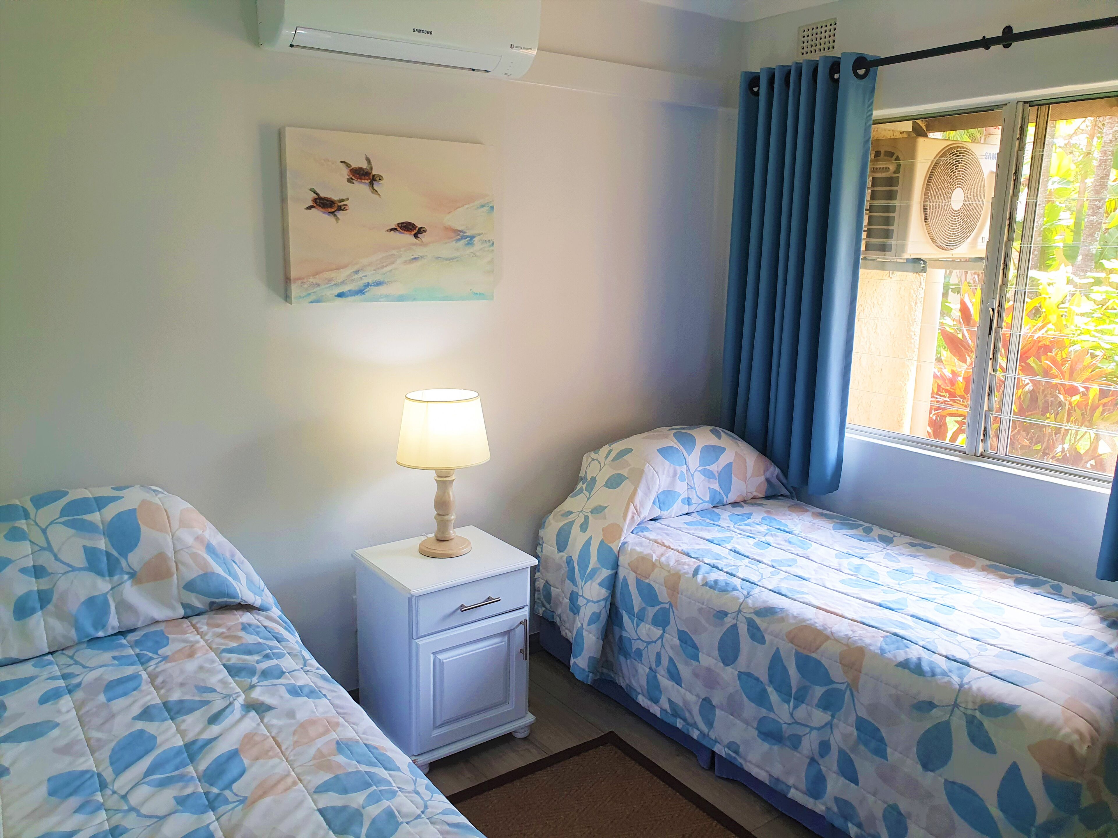 A room with two beds, a nightstand with a lamp, a painting on the wall, and a window with curtains.