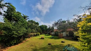 A well-maintained grassy area with a covered seating area, surrounded by lush greenery and a building with a red-tiled roof.