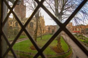 A view through a window with a geometric pattern, featuring a historic building with a large tower, a grassy area, and a paved path. - Coventry Historic Houses (Coventry)