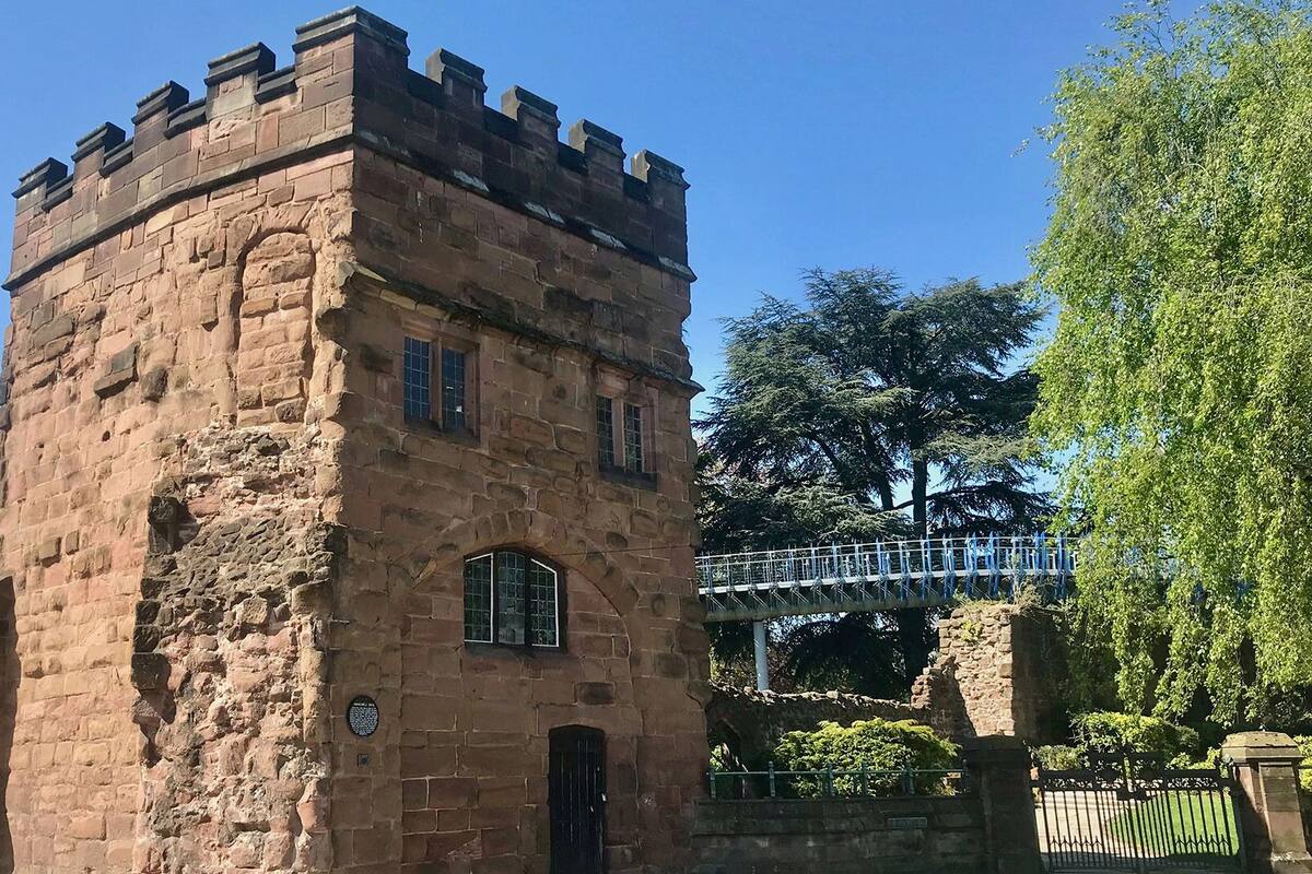 A historic stone tower with arched windows and a small door, surrounded by greenery and a pedestrian bridge.