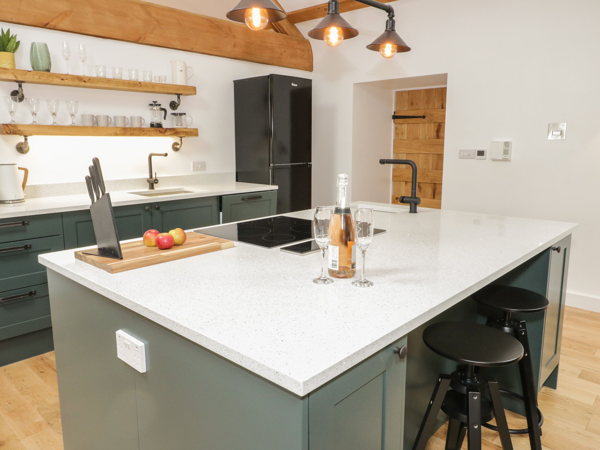 A modern kitchen with a large island, wooden shelves, and a black refrigerator.
