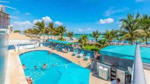 A pool area with lounge chairs and palm trees, a building with a sloped roof, and a clear blue sky. - Oceanview Apt at Marenas Resort, Sunny Isles Beach with Direct Beach Access!  (Sunny Isles Beach)