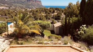 Paisagem costeira com vista para o mar e uma montanha distante. Há árvores, uma piscina e um edifício com telhado de azulejos.