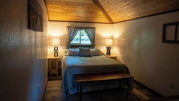 A bedroom with a sloped ceiling, a bed with a bench, two bedside lamps, and a window with curtains.