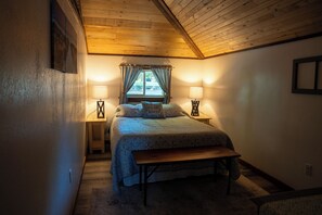 Une chambre avec un plafond en pente, un lit avec une banquette, deux lampes de chevet et une fenêtre avec des rideaux.
