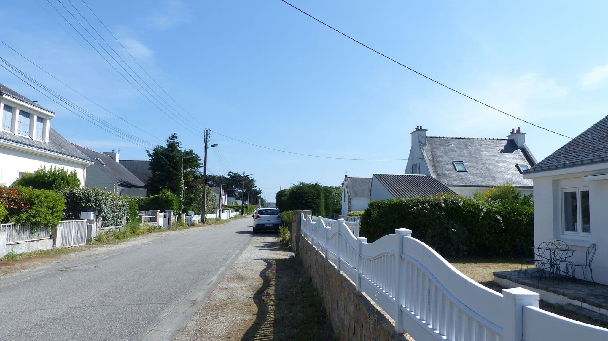 A narrow street lined with white houses, a parked car, and a white fence along the side.