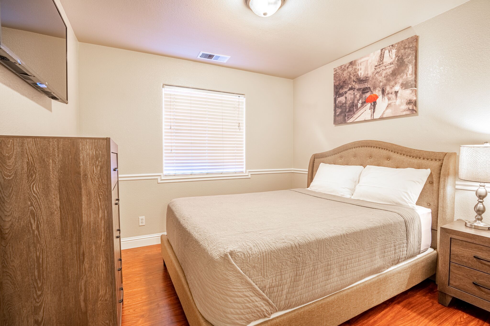 A bedroom with a bed, a nightstand, a dresser, a window with blinds, and a wall-mounted picture.