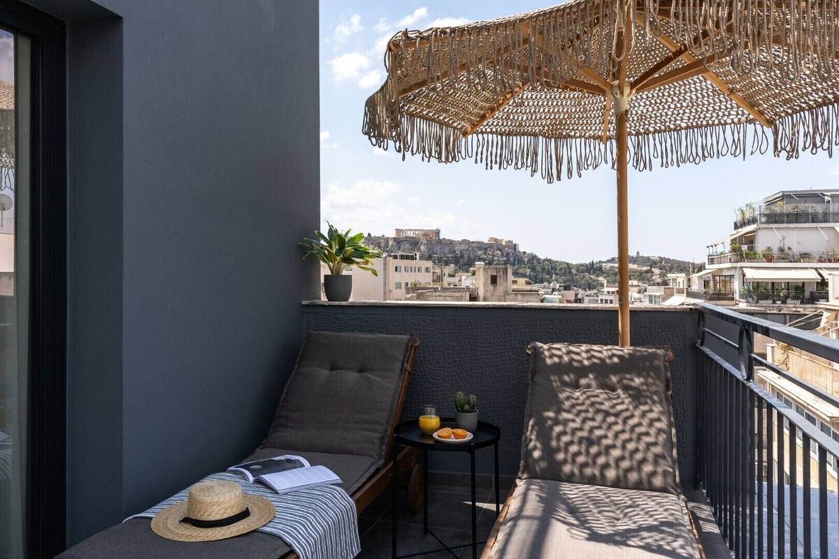 A balcony with a view of a cityscape, a woven umbrella, two lounge chairs, a small table with a plant and fruit, and a potted plant on the windowsill.