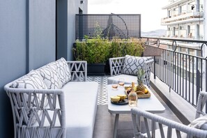 A balcony with a white sofa, a dining table with a bottle and glasses, and a view of a building across the street.