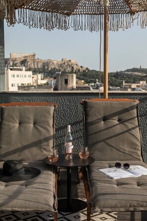 A rooftop terrace with two cushioned seating areas, a small table with a bottle and glasses, and a view of a historic building in the background.