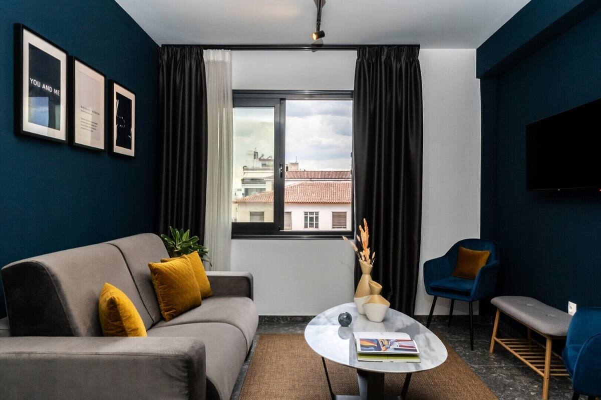 A modern living room with a grey sofa, a blue armchair, a round coffee table, and a view of buildings through a large window.