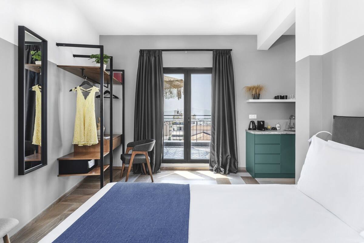 A modern hotel room with a bed, a desk with a chair, a wardrobe, a mirror, and a view of the outside balcony.