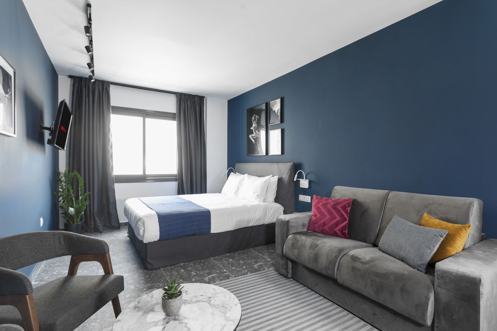 A modern hotel room with a bed, grey sofa, armchair, and a small round table with a plant.