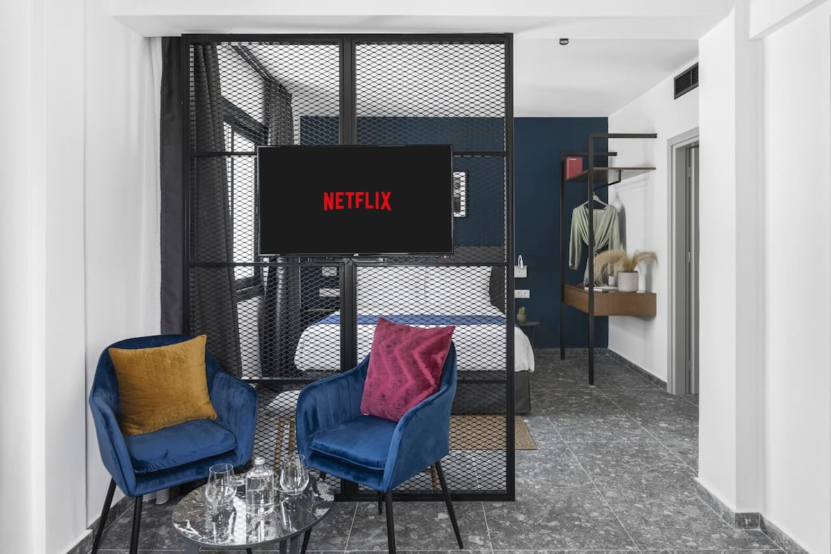 A modern hotel room with a large TV screen displaying 'NETFLIX', two blue armchairs with cushions, a small round table with glasses, and a bed with a blue headboard.