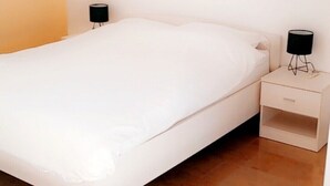 Ein Schlafzimmer mit einem weißen Bett, einem Nachttisch mit einer Lampe und einer schlichten Wand.
