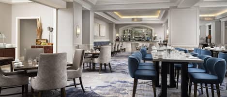 A spacious hotel dining area with multiple tables and chairs, a central chandelier, and a modern interior design.